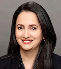 Professional headshot of Dr Brar smiling with long dark hair, wearing a black blazer against a neutral gray background.